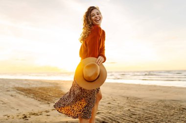 Gün batımında sahilde güzel bir kadının portresi. Kız plaj boyunca yürüyor ve sıcak sonbahar havasının tadını çıkarıyor. Seyahat, hafta sonu, rahatlama ve yaşam tarzı konsepti.