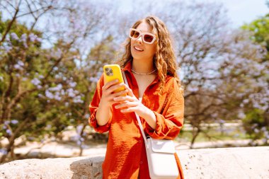 Genç bir kadın akıllı telefon kullanıyor ve şehirde yürürken gülümsüyor. İş, teknoloji, blog, iletişim kavramı.