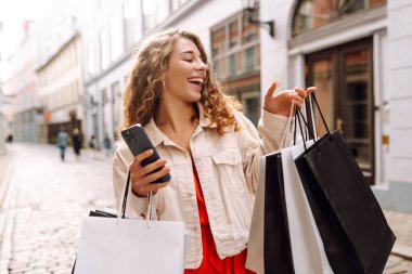 Güzel bir kadın alışveriş torbalarını açık hava mağazasında mutluluk içinde tutarken cep telefonu taksitlerini kullanıyor. Mobil uygulamalar kullanan bir bayan dijital mağazadan moda indirimi giysileri satın alıyor. Tüketim, Kara Cuma. İnternetten alışveriş.