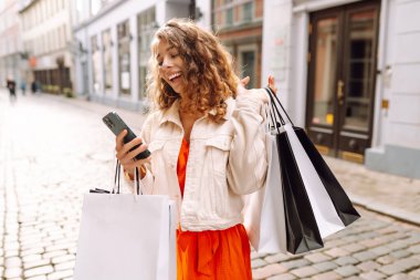 Güzel bir kadın alışveriş torbalarını açık hava mağazasında mutluluk içinde tutarken cep telefonu taksitlerini kullanıyor. Mobil uygulamalar kullanan bir bayan dijital mağazadan moda indirimi giysileri satın alıyor. Tüketim, Kara Cuma. İnternetten alışveriş.