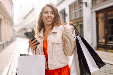 Güzel bir kadın alışveriş torbalarını açık hava mağazasında mutluluk içinde tutarken cep telefonu taksitlerini kullanıyor. Mobil uygulamalar kullanan bir bayan dijital mağazadan moda indirimi giysileri satın alıyor. Tüketim, Kara Cuma. İnternetten alışveriş.