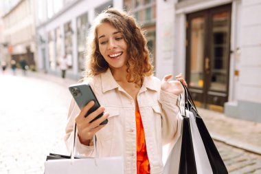 Pretty woman using instant mobile payments while holding shopping bags in the outdoor department store with happiness. Lady using mobile apps buy fashion sale clothes online in a digital store. Consumerism, black Friday. Online shopping.