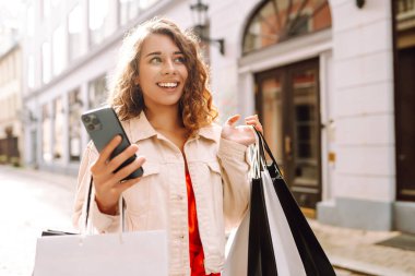 Güzel bir kadın alışveriş torbalarını açık hava mağazasında mutluluk içinde tutarken cep telefonu taksitlerini kullanıyor. Mobil uygulamalar kullanan bir bayan dijital mağazadan moda indirimi giysileri satın alıyor. Tüketim, Kara Cuma. İnternetten alışveriş.