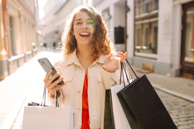 Güzel bir kadın alışveriş torbalarını açık hava mağazasında mutluluk içinde tutarken cep telefonu taksitlerini kullanıyor. Mobil uygulamalar kullanan bir bayan dijital mağazadan moda indirimi giysileri satın alıyor. Tüketim, Kara Cuma. İnternetten alışveriş.