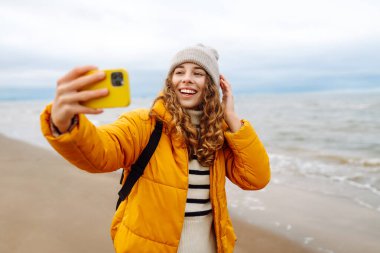 Okyanusta telefonu olan sarı ceketli mutlu bir kadın. Selfie zamanı. Soğuk rüzgarlı hava. Açık havada. Seyahat kavramı.