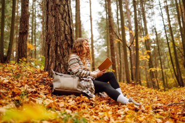 Hoş bir kadın, sonbahar sarı parkında bir ağacın altında oturmuş kitap okuyor. Eğitim. Doğadan zevk alma kavramı.