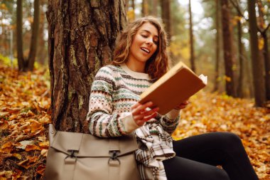 Hoş bir kadın, sonbahar sarı parkında bir ağacın altında oturmuş kitap okuyor. Eğitim. Doğadan zevk alma kavramı.