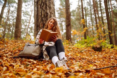 Hoş bir kadın, sonbahar sarı parkında bir ağacın altında oturmuş kitap okuyor. Eğitim. Doğadan zevk alma kavramı.