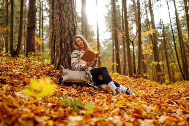 Hoş bir kadın, sonbahar sarı parkında bir ağacın altında oturmuş kitap okuyor. Eğitim. Doğadan zevk alma kavramı.