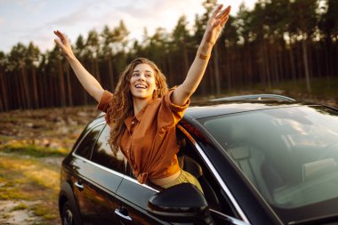 Genç bir kadın araba kullanırken pencereden dışarı bakarak taze rüzgarın tadını çıkarıyor. Yaşam tarzı, seyahat, turizm, doğa, aktif yaşam. Araba yolculuğu.