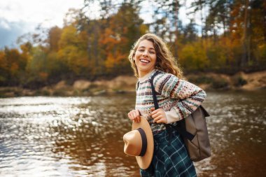 Sırt çantalı mutlu kadın gezgin nehir boyunca sonbahar yürüyüşünün tadını çıkarıyor. Aktif yaşam tarzı. Yürüyüş, seyahat, tatil konsepti.