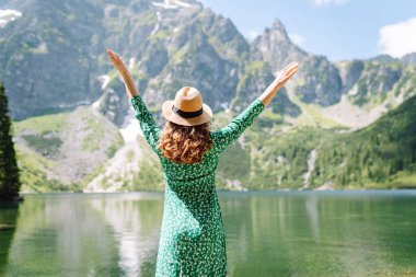 Parlak yeşil elbiseli ve şapkalı güzel bir turist kıyı gölünde yürüyor ve dağ manzarasının tadını çıkarıyor. Seyahat blogu. Macera, tatil konsepti.