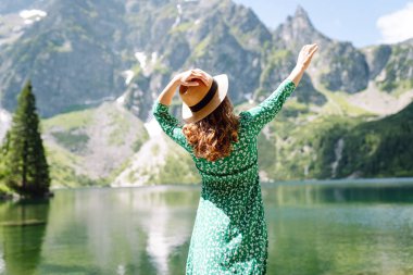 Parlak yeşil elbiseli ve şapkalı güzel bir turist kıyı gölünde yürüyor ve dağ manzarasının tadını çıkarıyor. Seyahat blogu. Macera, tatil konsepti.