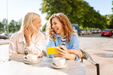 Yazlık kafede otururken mutlu kadınlar kahve içip cep telefonu kullanıyor. Moda, güzellik, blog, turizm.