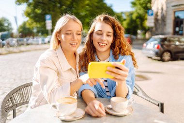 Yazlık kafede otururken mutlu kadınlar kahve içip cep telefonu kullanıyor. Moda, güzellik, blog, turizm.