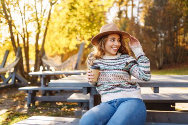 Sonbahar parkında elinde kahveyle yürüyen çekici, genç bir kadın. Seyahat, kamp, doğa konsepti