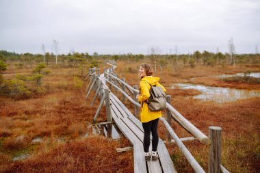 Sırt çantalı mutlu bir kadın bataklıkta tahta bir patika boyunca yürüyor. Seyahat konsepti, keşif. Etkin yaşam biçimi.