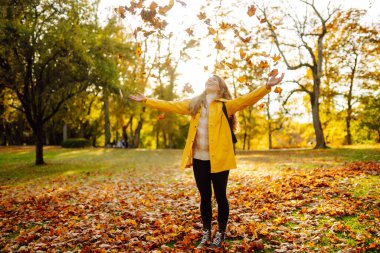 Elleri havada gülümseyen genç bir kadın ormandaki sonbahar yapraklarını fırlatıyor. Güzel bir yer. Rahatlama, eğlence, doğa ile yalnızlık kavramı..