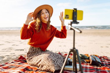 Dengeleyicili akıllı telefon kullanan, fotoğraf çeken ve deniz sahilinde canlı yayın yapan mutlu bir kadın. Mutlu kadın içerik yaratır. Canlı oturum günlüğü.