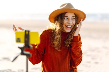 Dengeleyicili akıllı telefon kullanan, fotoğraf çeken ve deniz sahilinde canlı yayın yapan mutlu bir kadın. Mutlu kadın içerik yaratır. Canlı oturum günlüğü.