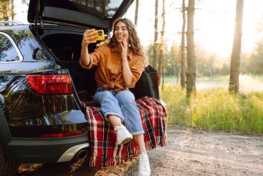 Güzel bir kadın arabanın bagajında dinleniyor ve selfie çekiyor. Seyahat, hafta sonu, doğa, rahatlama ve yaşam tarzı konsepti