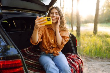 Güzel bir kadın arabanın bagajında dinleniyor ve selfie çekiyor. Seyahat, hafta sonu, doğa, rahatlama ve yaşam tarzı konsepti