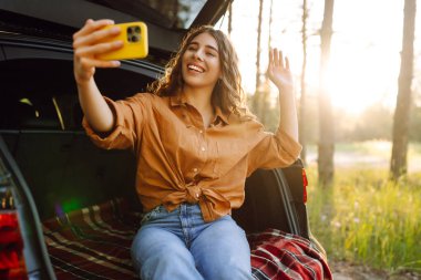 Güzel bir kadın arabanın bagajında dinleniyor ve selfie çekiyor. Seyahat, hafta sonu, doğa, rahatlama ve yaşam tarzı konsepti