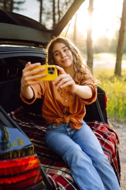 Güzel bir kadın arabanın bagajında dinleniyor ve selfie çekiyor. Seyahat, hafta sonu, doğa, rahatlama ve yaşam tarzı konsepti