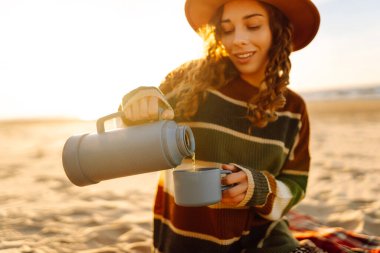 Şirin bir kadın sahilde oturup termostan sıcak bir içecek içiyor. Piknik, hafta sonu, rahatlama ve yaşam tarzı konsepti