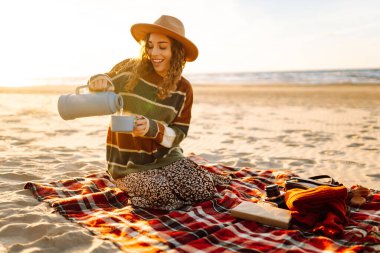 Şirin bir kadın sahilde oturup termostan sıcak bir içecek içiyor. Piknik, hafta sonu, rahatlama ve yaşam tarzı konsepti
