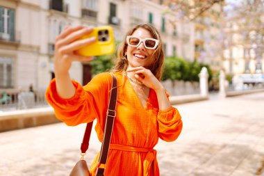 Kameraya poz veren mutlu, çekici genç bir kadın güneş ışığında selfie 'yi gülümsetiyor. Seyahat, turizm, teknoloji, blog, iletişim kavramı.