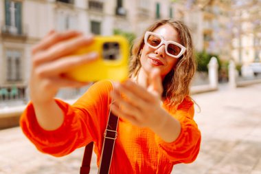 Kameraya poz veren mutlu, çekici genç bir kadın güneş ışığında selfie 'yi gülümsetiyor. Seyahat, turizm, teknoloji, blog, iletişim kavramı.