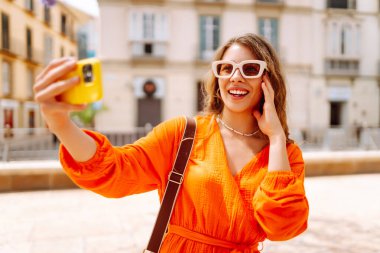 Kameraya poz veren mutlu, çekici genç bir kadın güneş ışığında selfie 'yi gülümsetiyor. Seyahat, turizm, teknoloji, blog, iletişim kavramı.