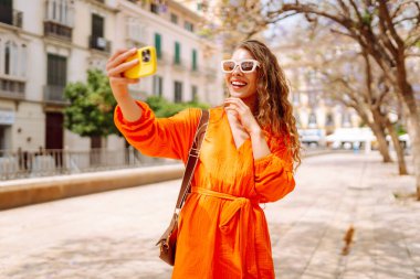 Kameraya poz veren mutlu, çekici genç bir kadın güneş ışığında selfie 'yi gülümsetiyor. Seyahat, turizm, teknoloji, blog, iletişim kavramı.