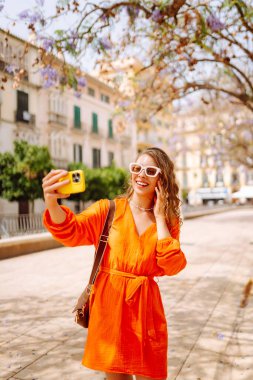 Kameraya poz veren mutlu, çekici genç bir kadın güneş ışığında selfie 'yi gülümsetiyor. Seyahat, turizm, teknoloji, blog, iletişim kavramı.
