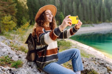 Mutlu kadın dinleniyor ve tatildeyken dağlarla ve akıllı telefondan gölle muhteşem manzaranın fotoğraflarını çekiyor. Macera, doğa, blog yazma.