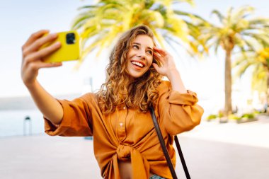 Genç bir kadın sokakta yürüyor ve akıllı telefon kamerasıyla selfie çekiyor. Teknoloji, blog yazma, iletişim kavramı.