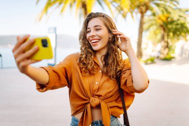 Genç bir kadın sokakta yürüyor ve akıllı telefon kamerasıyla selfie çekiyor. Teknoloji, blog yazma, iletişim kavramı.