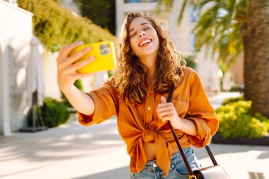 Genç bir kadın sokakta yürüyor ve akıllı telefon kamerasıyla selfie çekiyor. Teknoloji, blog yazma, iletişim kavramı.