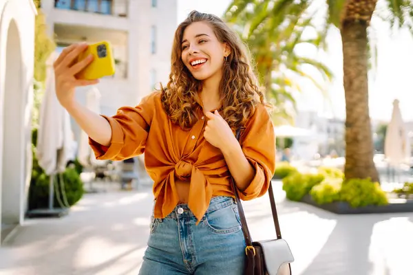 Genç bir kadın sokakta yürüyor ve akıllı telefon kamerasıyla selfie çekiyor. Teknoloji, blog yazma, iletişim kavramı.