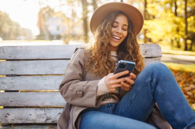 Sonbahar parkındaki bankta cep telefonu kullanan mutlu bir kadın, başvuru yaparken gülümseyen genç bir kadın. Tatil kavramı, blog yazma, teknoloji, hafta sonu.