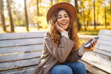 Sonbahar parkındaki bankta cep telefonu kullanan mutlu bir kadın, başvuru yaparken gülümseyen genç bir kadın. Tatil kavramı, blog yazma, teknoloji, hafta sonu.