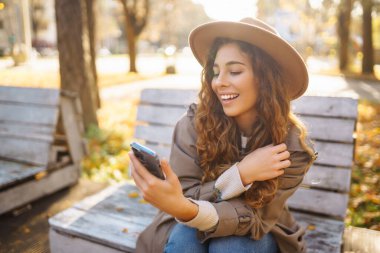 Sonbahar parkındaki bankta cep telefonu kullanan mutlu bir kadın, başvuru yaparken gülümseyen genç bir kadın. Tatil kavramı, blog yazma, teknoloji, hafta sonu.