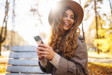 Sonbahar parkındaki bankta cep telefonu kullanan mutlu bir kadın, başvuru yaparken gülümseyen genç bir kadın. Tatil kavramı, blog yazma, teknoloji, hafta sonu.