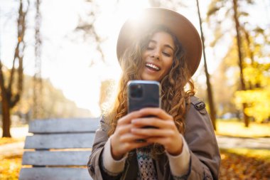 Sonbahar parkındaki bankta cep telefonu kullanan mutlu bir kadın, başvuru yaparken gülümseyen genç bir kadın. Tatil kavramı, blog yazma, teknoloji, hafta sonu.