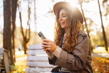 Sonbahar parkındaki bankta cep telefonu kullanan mutlu bir kadın, başvuru yaparken gülümseyen genç bir kadın. Tatil kavramı, blog yazma, teknoloji, hafta sonu.