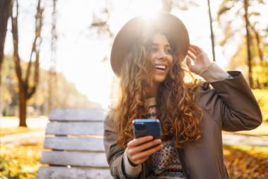 Sonbahar parkındaki bankta cep telefonu kullanan mutlu bir kadın, başvuru yaparken gülümseyen genç bir kadın. Tatil kavramı, blog yazma, teknoloji, hafta sonu.