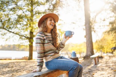 Gülümseyen genç kadın ormanda oturur, çay içer. Seyahatin, tatilin ve sıcak kahvenin tadını çıkar. Yürüyüş yaşam tarzı, macera ve turizm, dinlenme, rahatlama.