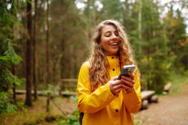 Akıllı telefon kullanan ve sonbahar havasının tadını çıkaran genç bir kadın. Sonbahar ormanında yürüyen bir kadın. Rahatlama, keyif, doğa ile yalnızlık. Bloglama, teknoloji.