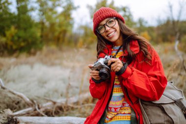 Kameralı ve sırt çantalı bir kadın sonbahar ormanında yürüyüş yapıyor. Doğayı fotoğraflayan dişi turist. Doğa fotoğrafçısı konsepti.
